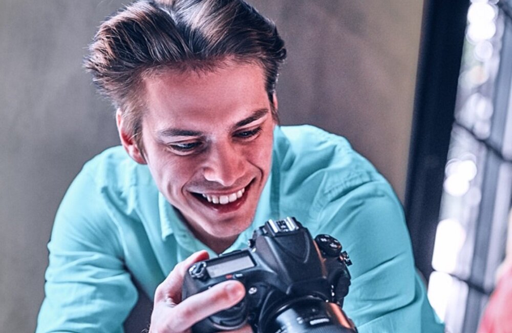 Osoba w jasnoniebieskiej koszuli trzyma aparat fotograficzny i patrzy na niego z uśmiechem.