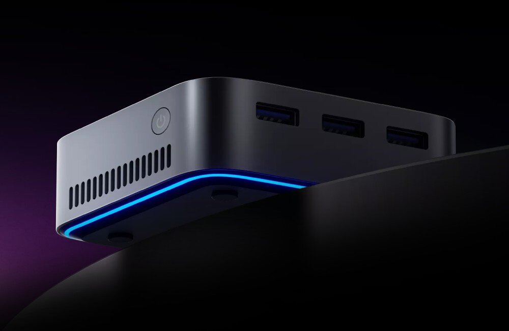 Na obrazie widoczny jest kompaktowy komputer w ciemnym kolorze z podświetleniem LED na krawędzi. Posiada kilka portów USB oraz przycisk zasilania na obudowie.