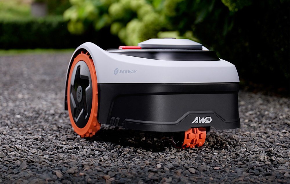 Robot koszący z logo \'SEGWAY\' i \'AWD\' na obudowie, stoi na żwirowej powierzchni. Urządzenie ma czarne i białe elementy z pomarańczowymi kołami.