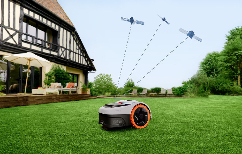 Na trawniku przed domem znajduje się robot koszący z pomarańczowymi kołami. W tle widać trzy satelity na niebie.