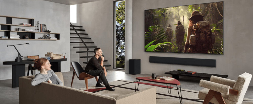 W nowoczesnym salonie na ścianie wisi duży telewizor, na którym wyświetlana jest scena z dżungli. W pomieszczeniu znajdują się meble, takie jak sofa, krzesła i stolik, a na półkach stoją książki i dekoracje.