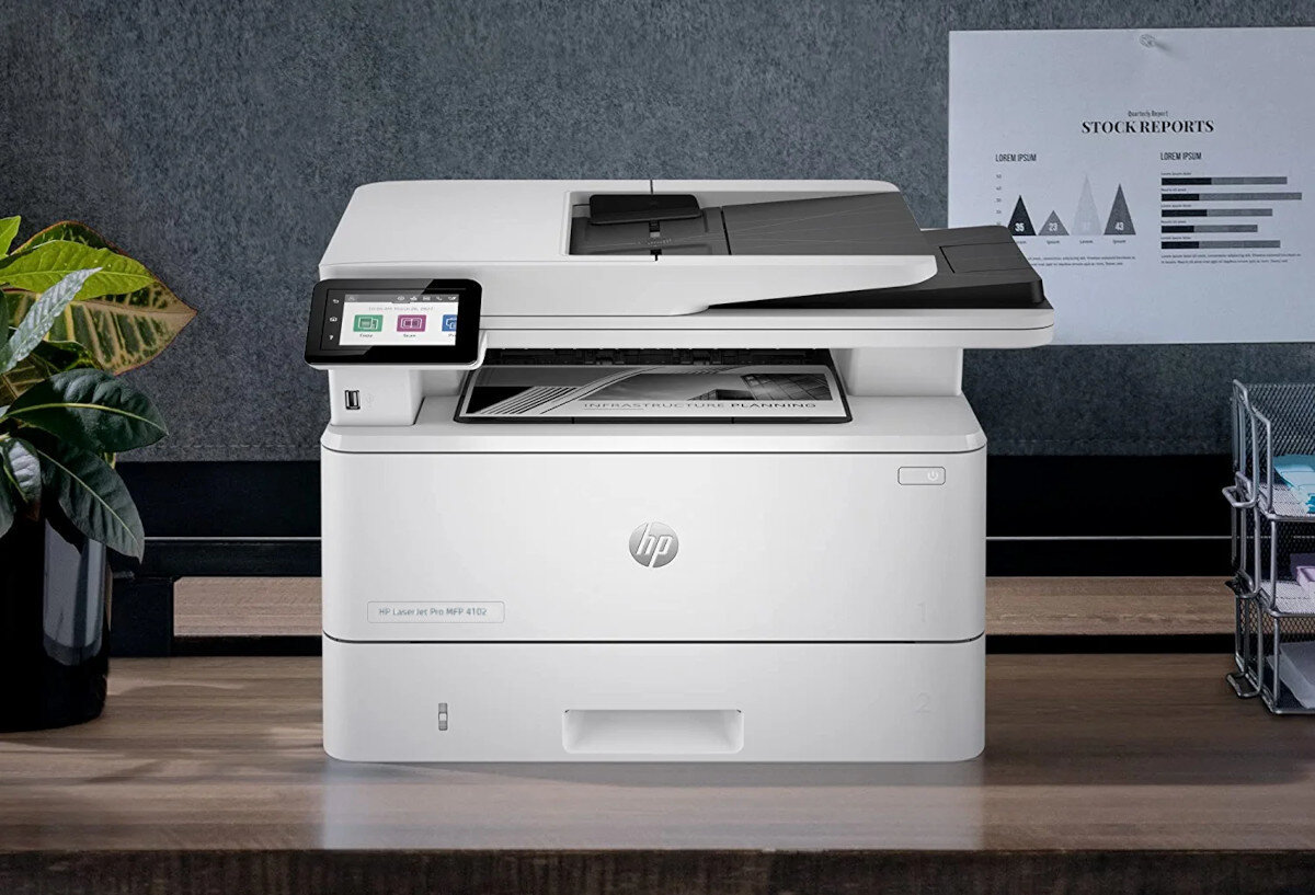 Na biurku stoi białe urządzenie wielofunkcyjne z logo HP, z wyświetlaczem dotykowym po lewej stronie. W tle znajduje się plakat z napisem \'STOCK REPORTS\' oraz wykresami.