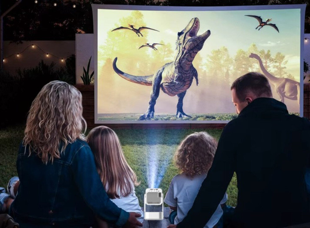 Grupa osób ogląda film z dinozaurami na dużym ekranie projekcyjnym na zewnątrz. Przed nimi znajduje się projektor emitujący światło na ekran.
