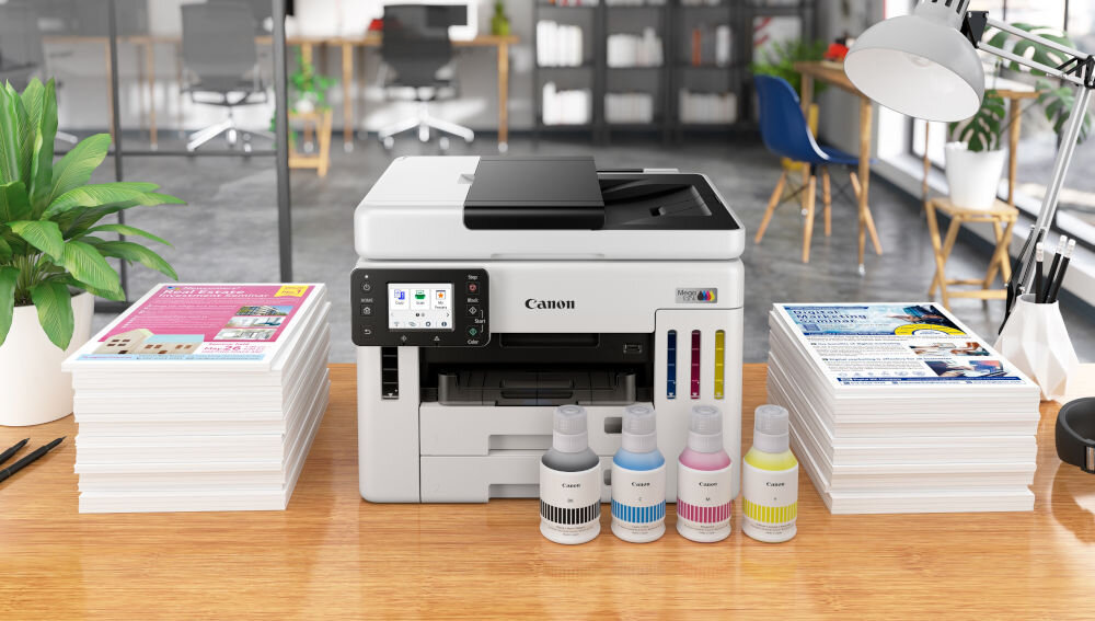 Na biurku znajduje się urządzenie wielofunkcyjne Canon z wyświetlaczem i logo producenta. Obok urządzenia stoją cztery butelki z tuszem oraz stosy papieru.