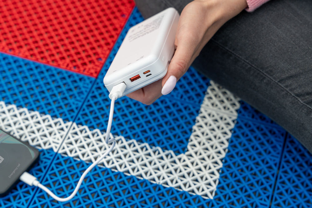 Osoba trzyma biały powerbank z podłączonym kablem, leżący na kolorowej, geometrycznej powierzchni. Obok znajduje się telefon również podłączony do powerbanka.