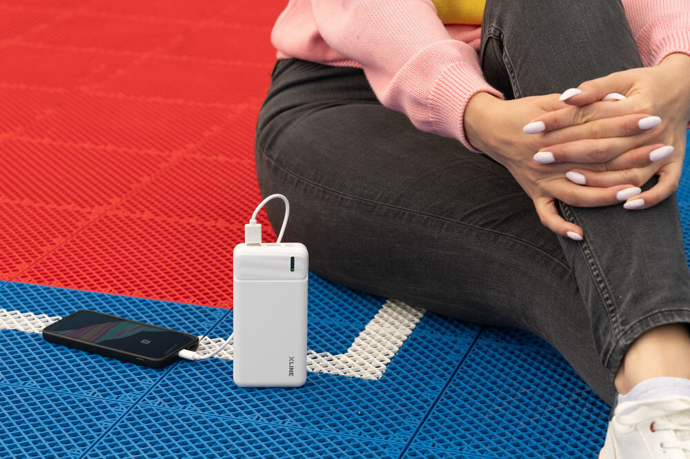 Osoba siedzi na kolorowej powierzchni, obok niej znajduje się biały powerbank z napisem \'XLINE\', podłączony do smartfona. Tło składa się z czerwonych i niebieskich paneli z białymi liniami.