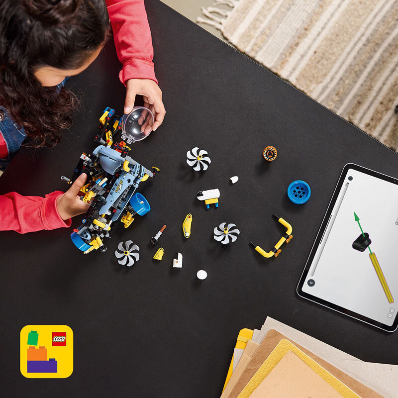Osoba składa model z klocków LEGO, otoczona różnymi elementami konstrukcyjnymi na czarnym stole. W rogu widoczny jest tablet z grafiką oraz logo LEGO w lewym dolnym rogu.