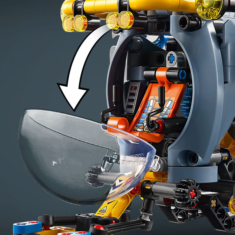 Zbliżenie na model LEGO przedstawiający wnętrze badawczej łodzi podwodnej z otwartą przezroczystą kopułą i widocznymi elementami konstrukcyjnymi w kolorach żółtym, pomarańczowym i niebieskim. Na górze znajduje się biała strzałka wskazująca kierunek otwierania kopuły.