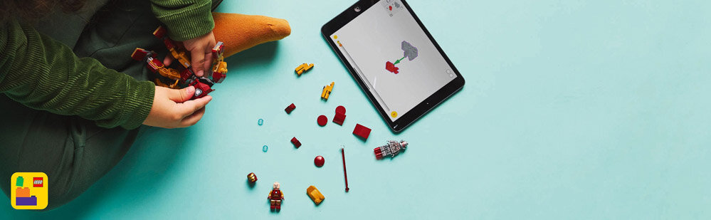 Osoba składa zestaw klocków LEGO, obok leży tablet z instrukcją budowy. Na podłodze rozłożone są różne elementy LEGO w kolorach czerwonym, żółtym i niebieskim.