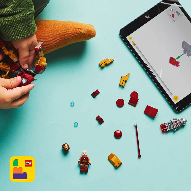 Na jasnoniebieskim tle znajdują się elementy zestawu LEGO, w tym figurki i klocki, obok tabletu z instrukcją. W lewym dolnym rogu widoczne jest logo LEGO.