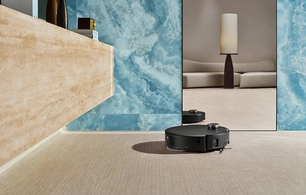 Czarny robot sprzątający porusza się po jasnej podłodze w eleganckim wnętrzu z niebieskimi ścianami. W tle widoczna jest sofa i lampa stojąca.