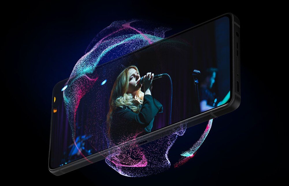 Smartfon wyświetla obraz kobiety śpiewającej do mikrofonu na scenie, otoczony kolorowymi efektami świetlnymi na czarnym tle.