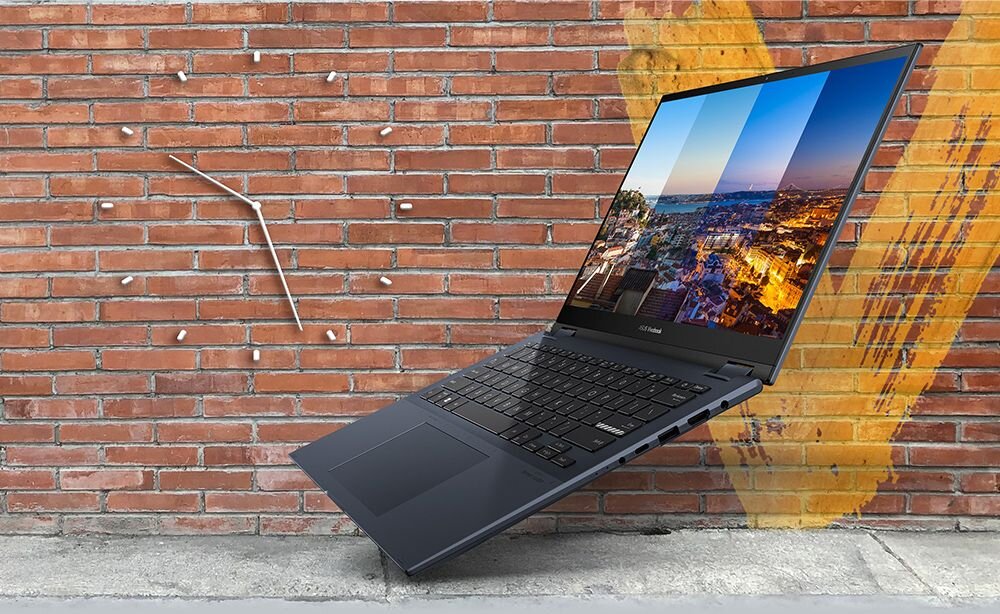 Laptop z otwartym ekranem wyświetlającym krajobraz miasta, umieszczony na tle ceglanej ściany z zegarem bez tarczy. Na ścianie widoczny jest duży, żółty znak w kształcie litery V.