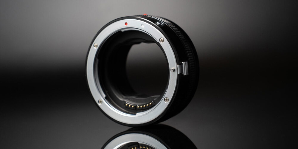 ADAPTER BAGNETOWY Z AUTOFOCUSEM MEGADAP EFTZ21 - CANON EF NIKON Z Metalowy adapter bagnetowy ustawiony pionowo na błyszczącej powierzchni, pokazany pod kątem na ciemnym, rozmytym tle