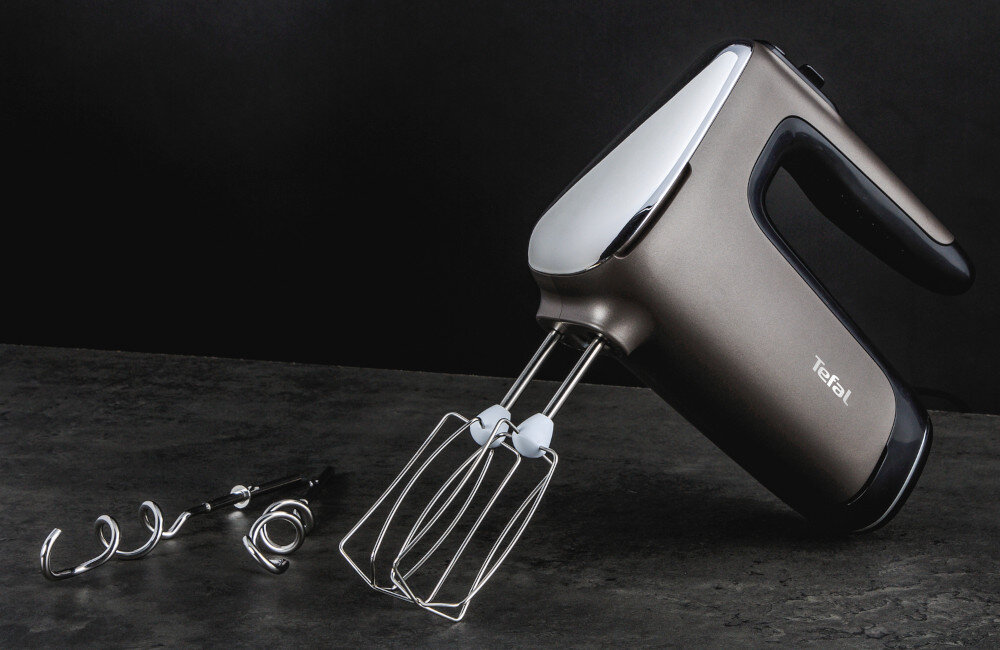 Na ciemnym tle znajduje się mikser ręczny z metalowymi końcówkami do mieszania i logo \'Tefal\' na obudowie. Obok leżą dwie spiralne końcówki do wyrabiania ciasta.