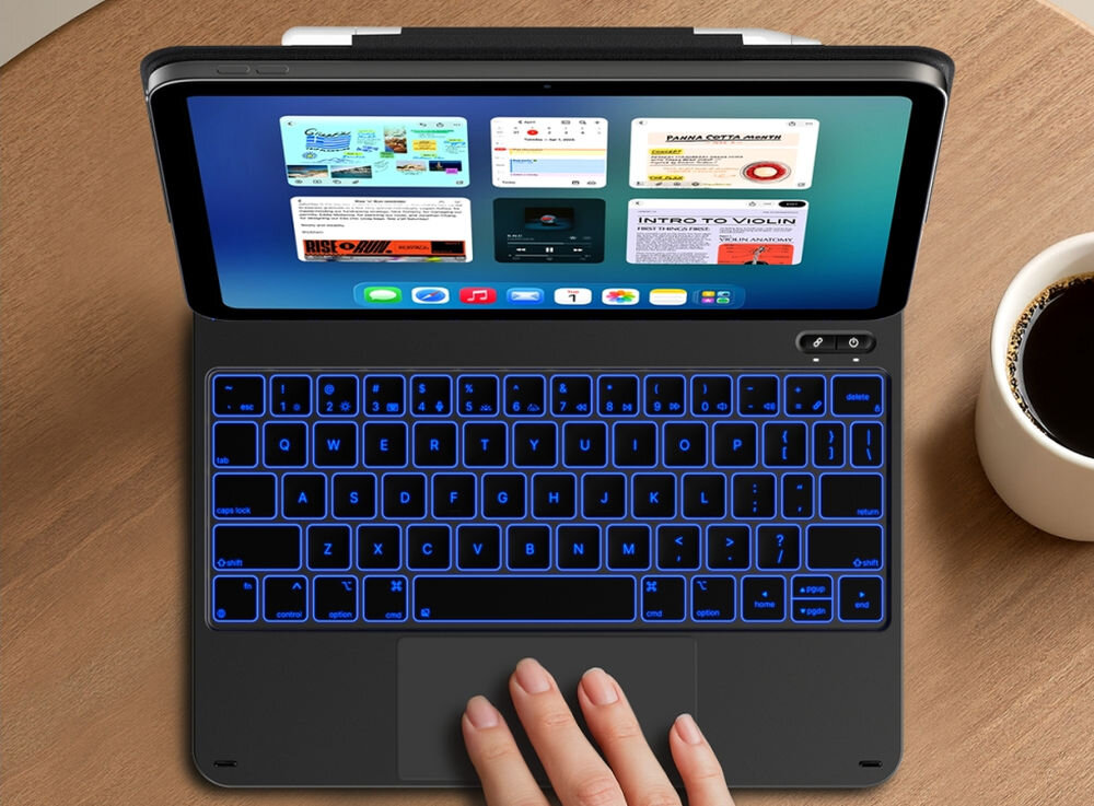 Na drewnianym stole znajduje się iPad z otwartą klawiaturą, na której widoczne są niebieskie podświetlane klawisze. Obok stoi filiżanka z czarną kawą.