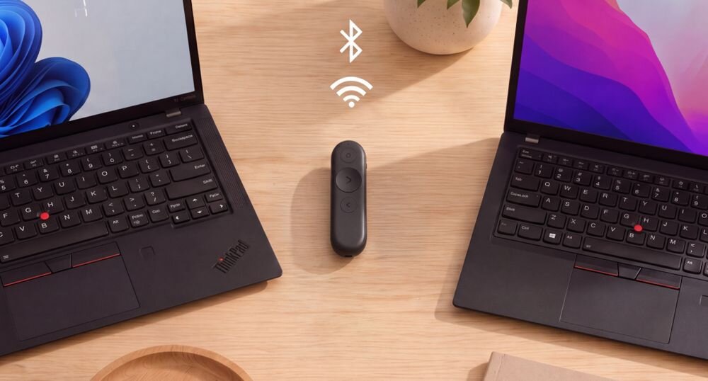 LENOVO Portable Rechargeable GXH1Q30399 Wbudowany akumulator  Pełne ładowanie 2,5 godziny 15 godzin Bluetooth 5.3  niska waga 18,8 g - Pilot do prezentacji leżący na drewnianym biurku pomiędzy dwoma otwartymi laptopami, z symbolami łączności bezprzewodowej
