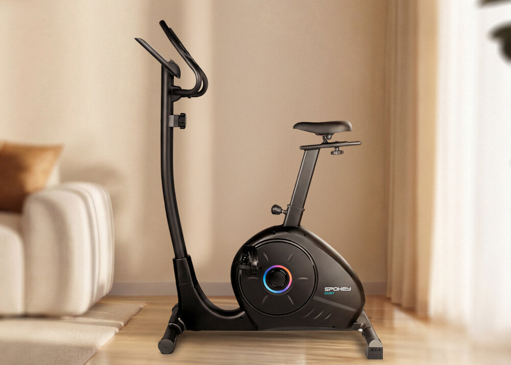 Na obrazie znajduje się czarny rower magnetyczny z napisem \'SPOKEY Dust\' na obudowie. Rower stoi w jasnym pomieszczeniu z drewnianą podłogą i zasłonami w tle.