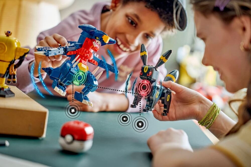 Dwie osoby bawią się zestawem klocków przedstawiającym postacie przypominające Pokémony. Na stole leży czerwono-biały przedmiot przypominający Pokéball.