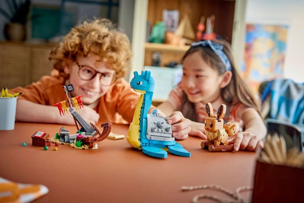 Na stole znajdują się klocki LEGO przedstawiające niebieskiego stwora z żółtymi elementami oraz brązowego stwora z dużymi uszami. W tle widać dwoje dzieci bawiących się tymi klockami.