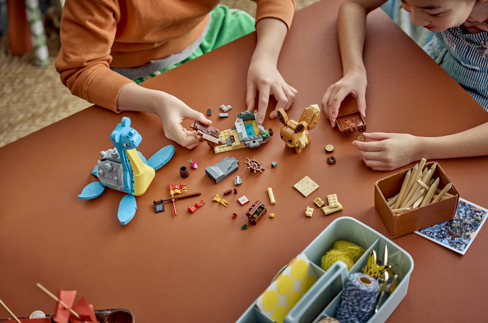 Na stole znajdują się klocki LEGO przedstawiające postacie i elementy związane z Pokémon, w tym Eevee i Lapras. Obok klocków widać ręce dzieci bawiących się zestawem oraz pojemniki z przyborami do rysowania.