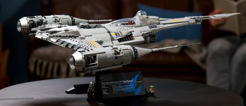 Модель космічного корабля LEGO Star Wars стоїть на темному столі. Перед моделлю висить табличка з написом «Зоряний винищувач N-1 Мандалорця».