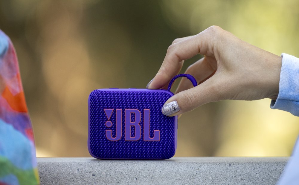 Fioletowy głośnik mobilny z widocznym logo \'JBL\' trzymany jest w dłoni. W tle rozmyte kolory natury.