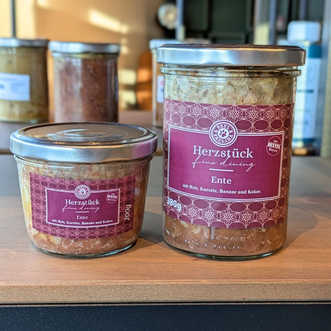 Na zdjęciu widoczne są dwa słoiki z etykietą \'Herzstück fine dining Ente mit Reis, Karotte, Banane und Kokos\'. Słoiki stoją na drewnianej powierzchni, a w tle widać inne słoiki.