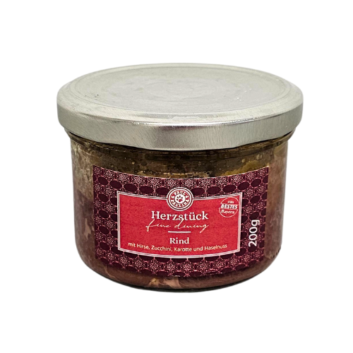 Słoik z metalową pokrywką zawiera produkt spożywczy z etykietą w kolorze czerwonym. Na etykiecie widnieje tekst: \'Herzstück feine Länderey Rind mit Hirse, Zucchini, Karotte und Haselnuss 200g\'.