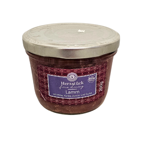 Słoik z metalową pokrywką zawiera produkt spożywczy, na etykiecie widnieje napis: \'Herzstück fein & fertig Lamm mit Hirse, Kürbis, Zucchini und Gurke\'.