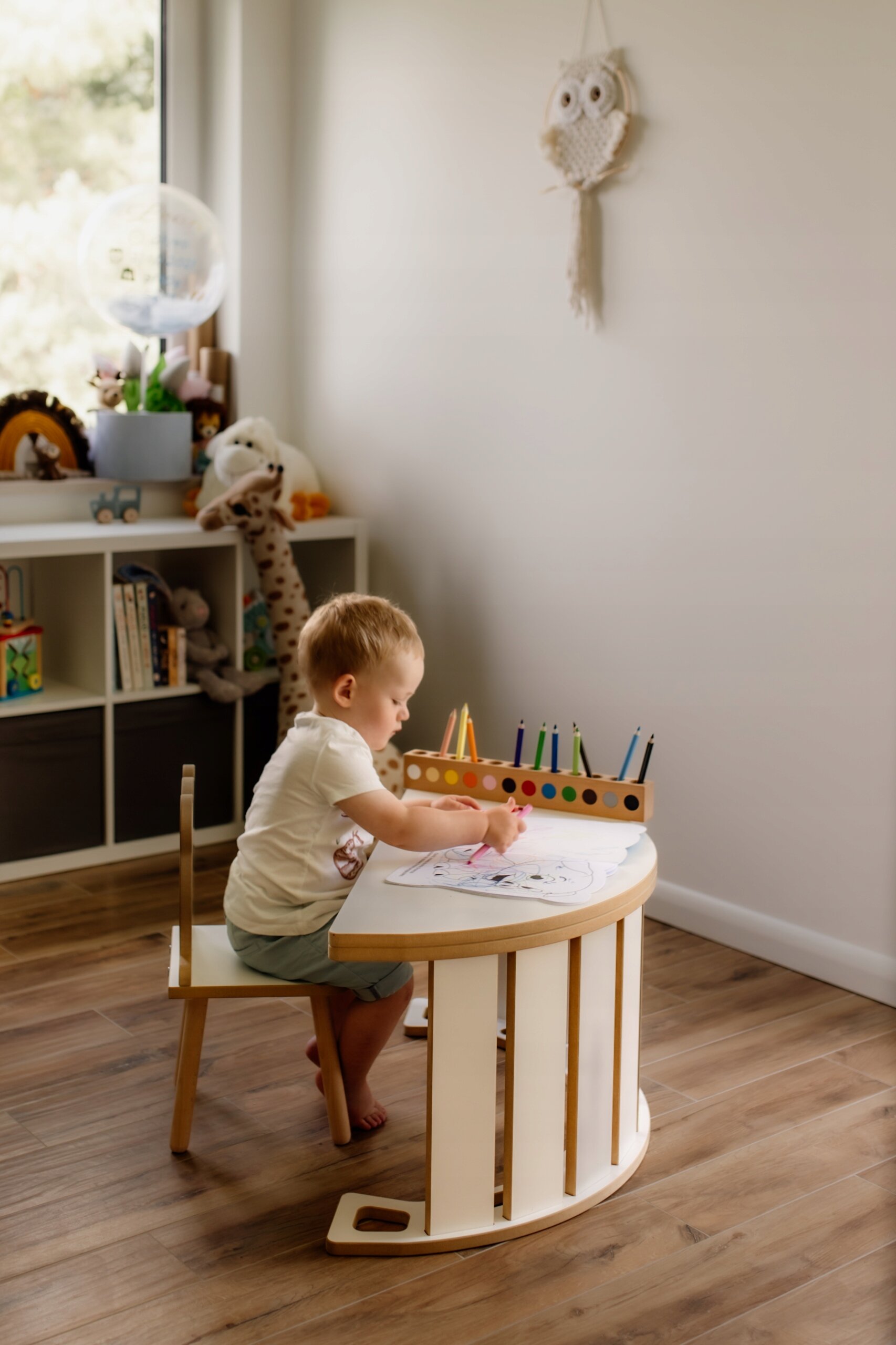 Dziecko siedzi przy białym bujaku Montessori, rysując na kartce papieru. W tle widać półkę z zabawkami i książkami oraz dekorację w kształcie sowy na ścianie.