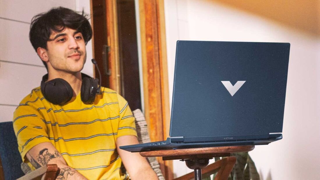 Osoba siedzi przy laptopie z widocznym logo w kształcie litery \'V\'. Laptop stoi na drewnianym stoliku, a osoba ma na szyi słuchawki.
