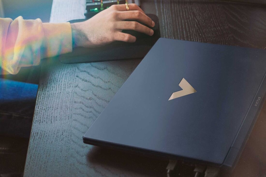 Na ciemnym drewnianym biurku leży zamknięty laptop z widocznym logo w kształcie litery \'V\'. Obok laptopa znajduje się ręka na myszce komputerowej.