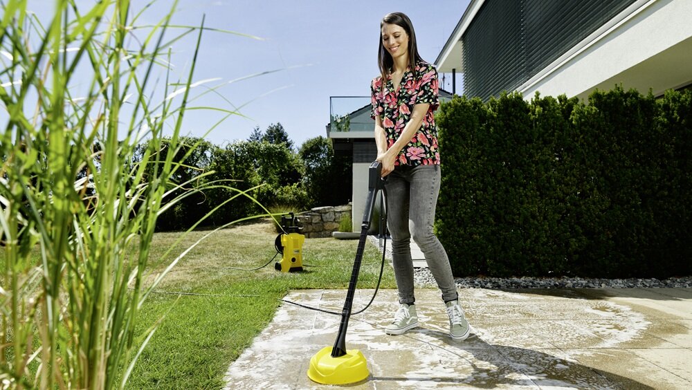 Osoba używa myjki ciśnieniowej z żółtą końcówką do czyszczenia powierzchni na zewnątrz budynku. W tle widoczny jest ogród z trawnikiem i krzewami.