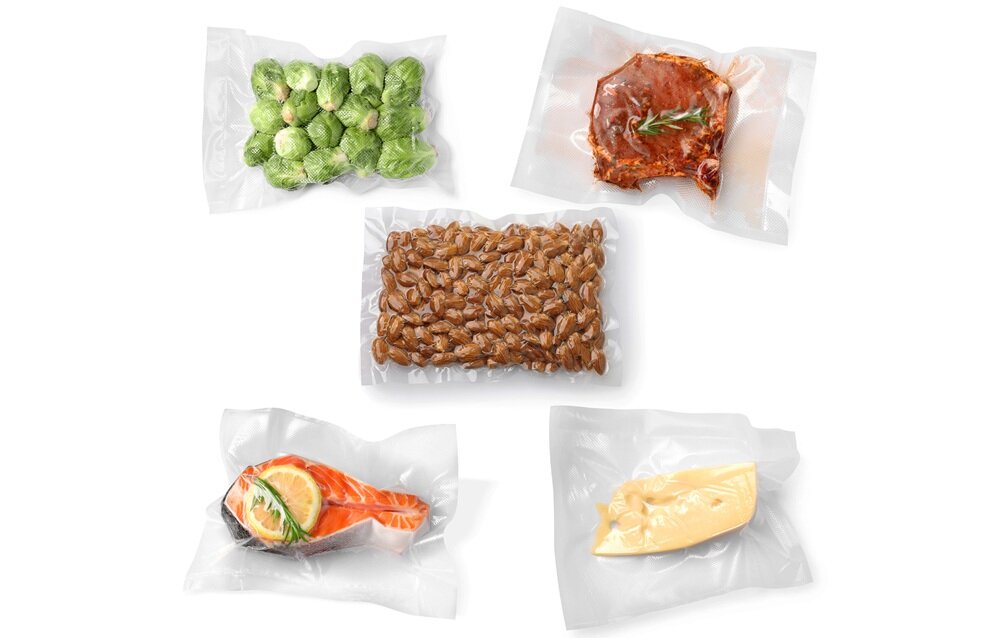 Na obrazie widoczne są różne produkty spożywcze zapakowane próżniowo w przezroczyste woreczki. Wśród nich znajdują się brukselki, kawałek mięsa, orzechy, kawałek łososia z cytryną oraz ser.
