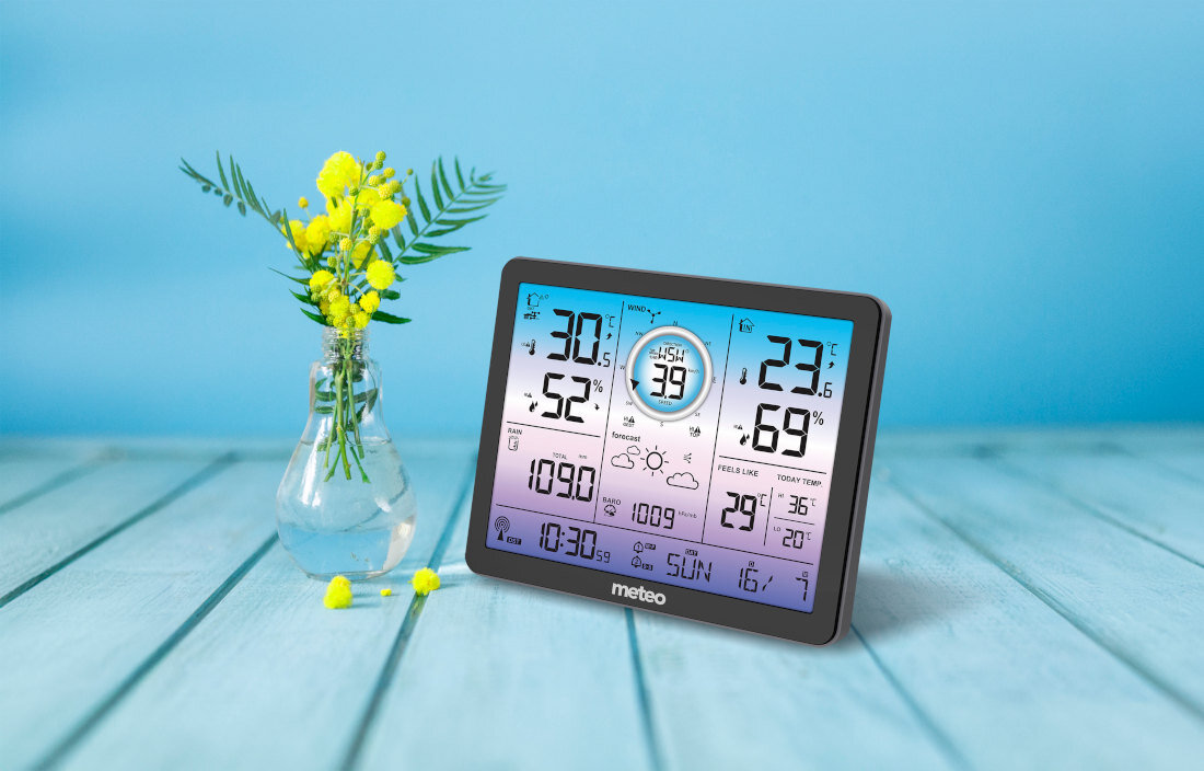 Stacja pogody z wyświetlaczem pokazującym temperaturę, wilgotność, ciśnienie i czas, z logo \'meteo\' na dole. Obok znajduje się wazon z żółtymi kwiatami na drewnianym stole, na tle niebieskiej ściany.