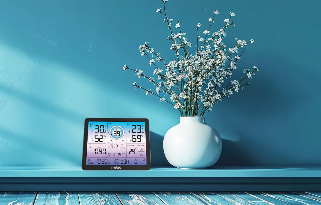 Na niebieskiej półce stoi stacja pogody z wyświetlaczem pokazującym różne dane meteorologiczne oraz biały wazon z gałązkami kwiatów. Na stacji widoczny jest napis \'meteo\'.