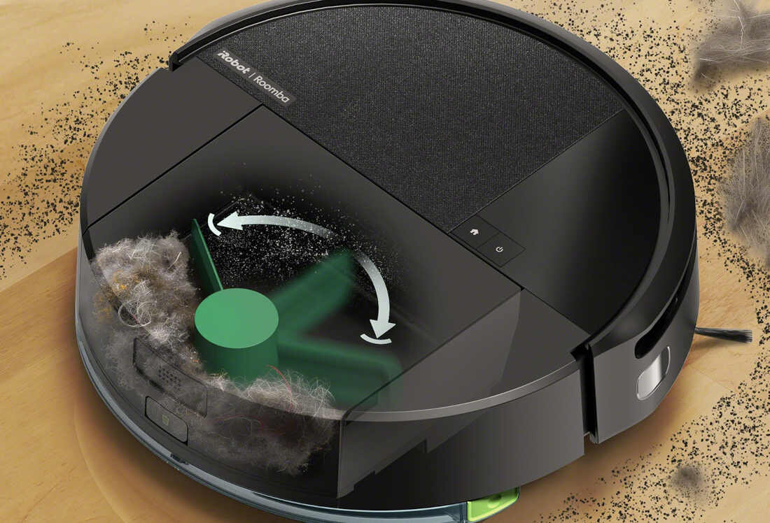 Czarny robot sprzątający z widocznym logo \'iRobot Roomba\' na górnej części obudowy, otoczony kurzem i sierścią. Wewnątrz urządzenia widoczna jest zielona szczotka z białymi strzałkami wskazującymi kierunek obrotu.