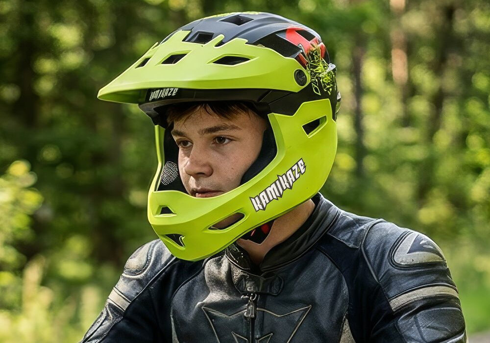 Osoba nosi zielony kask full face z napisem \'KAMIKAZE\' na tle zielonego lasu. Kask ma czarne i czerwone elementy oraz otwory wentylacyjne.