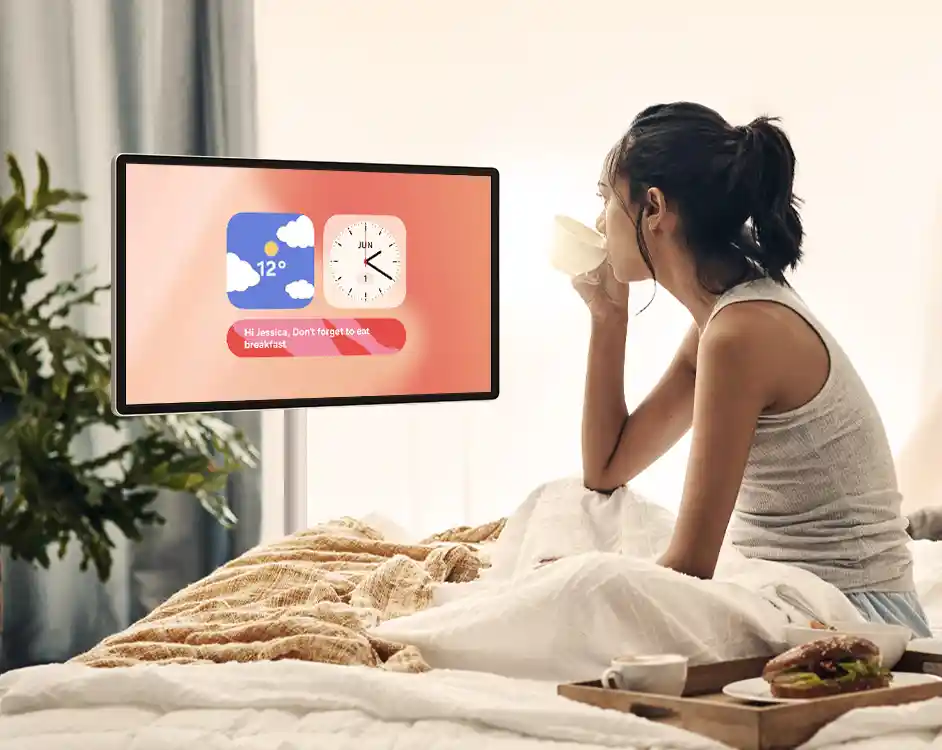 Osoba siedzi na łóżku, pijąc z kubka, obok znajduje się taca z jedzeniem. Na ekranie telewizora wyświetlane są ikony pogody i zegara oraz tekst: \'Hi Jessica, Don\'t forget to eat breakfast\'.