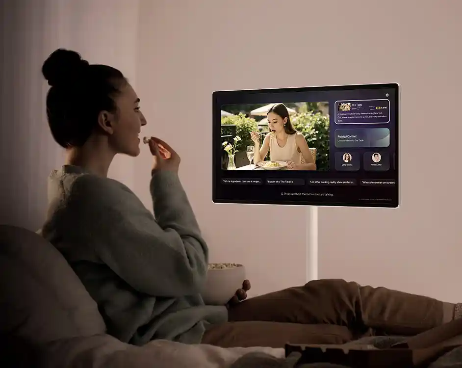 Osoba siedzi na łóżku z miską popcornu, oglądając telewizor. Na ekranie telewizora wyświetlana jest scena z filmu lub programu, przedstawiająca kobietę jedzącą przy stole.