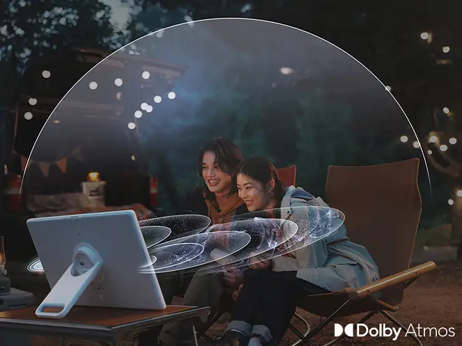 Dwie osoby siedzą na zewnątrz przed telewizorem, na którym widoczny jest efekt dźwięku przestrzennego. W prawym dolnym rogu znajduje się logo \'Dolby Atmos\'.