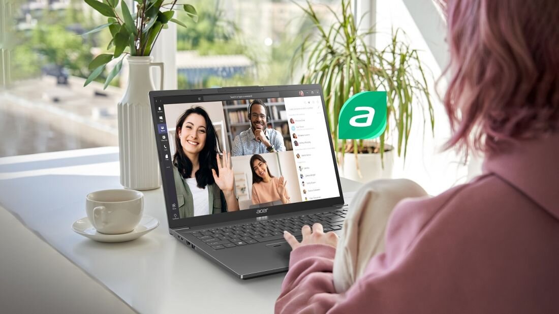 Osoba siedzi przy biurku z laptopem marki Acer, na którym widoczny jest ekran z wideokonferencją. Obok laptopa stoi filiżanka i wazon z rośliną, a w tle znajduje się okno z widokiem na zewnątrz.