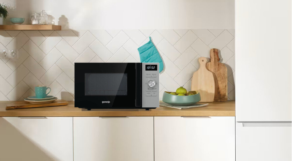 KUCHENKA MIKROFALOWA GORENJE MO20A4XH Kuchenka mikrofalowa Gorenje stoi na drewnianym blacie na tle jasnej ściany z białymi płytkami ułożonymi w jodełkę. Obok znajdują się deski do krojenia, miska z owocami, kubek oraz turkusowa rękawica kuchenna zawieszona na ścianie, a na panelu urządzenia widoczny jest czas 13:52 i pokrętło sterowania
