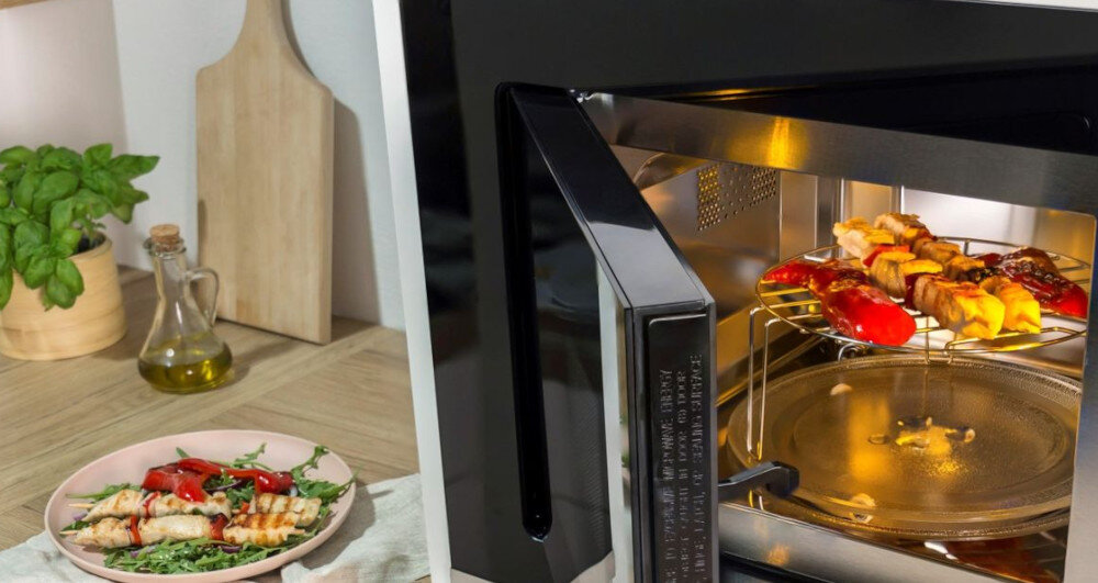 KUCHENKA MIKROFALOWA GORENJE MO20A4XH Otwarta kuchenka mikrofalowa z podświetlonym wnętrzem, w której na metalowej kratce podgrzewane są szaszłyki i warzywa. Obok na blacie stoi talerz z przygotowanym daniem, a w tle widoczne są drewniane akcesoria kuchenne, oliwa i roślina doniczkowa grill mikrofalówka chrupiące potrawy opiekanie przyrumienianie