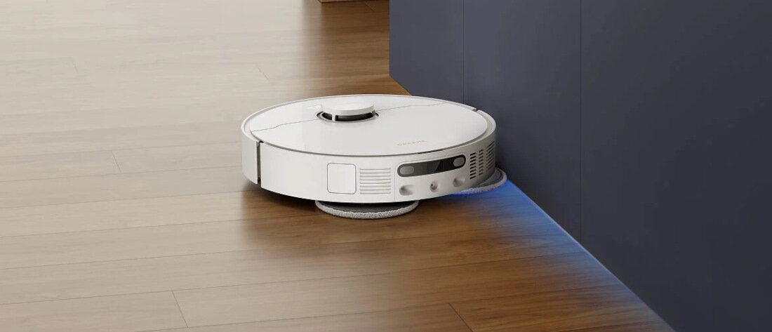 Na drewnianej podłodze znajduje się biały robot sprzątający, który przylega do ciemnej ściany. Robot ma okrągły kształt i widoczne są na nim przyciski oraz czujniki.