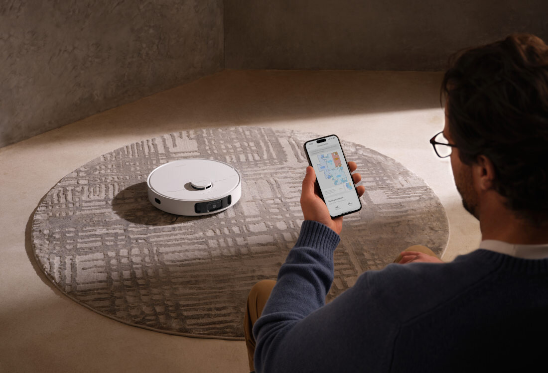 Osoba siedzi na podłodze, trzymając smartfon z wyświetloną aplikacją, podczas gdy na dywanie znajduje się biały robot sprzątający. Ściany i podłoga są w neutralnych kolorach.