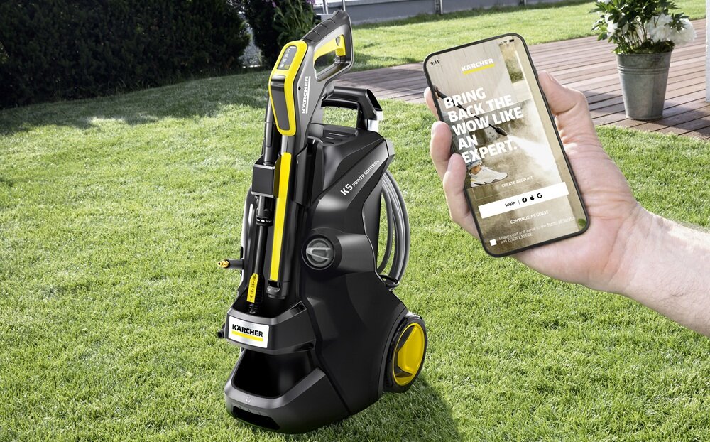 Na trawniku stoi czarna myjka ciśnieniowa z żółtymi elementami i widocznym logo KARCHER. Obok osoba trzyma smartfon z aplikacją, na której widnieje tekst: \'BRING BACK THE WOW LIKE AN EXPERT.\'