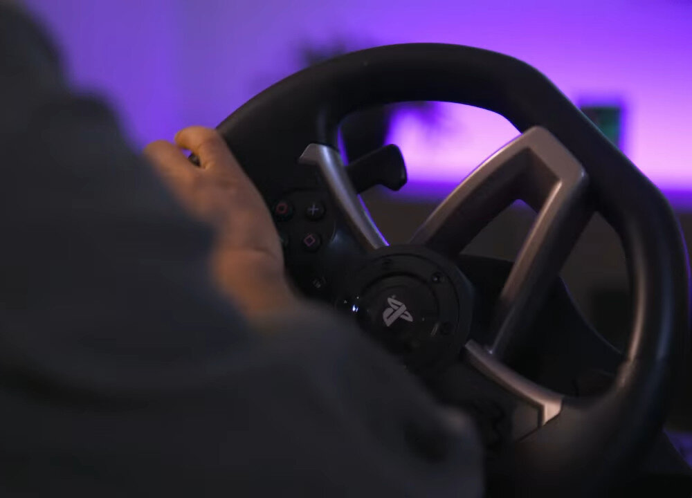 Osoba trzyma kierownicę do gier z widocznym logo PlayStation na środku. W tle widoczne jest fioletowe oświetlenie.