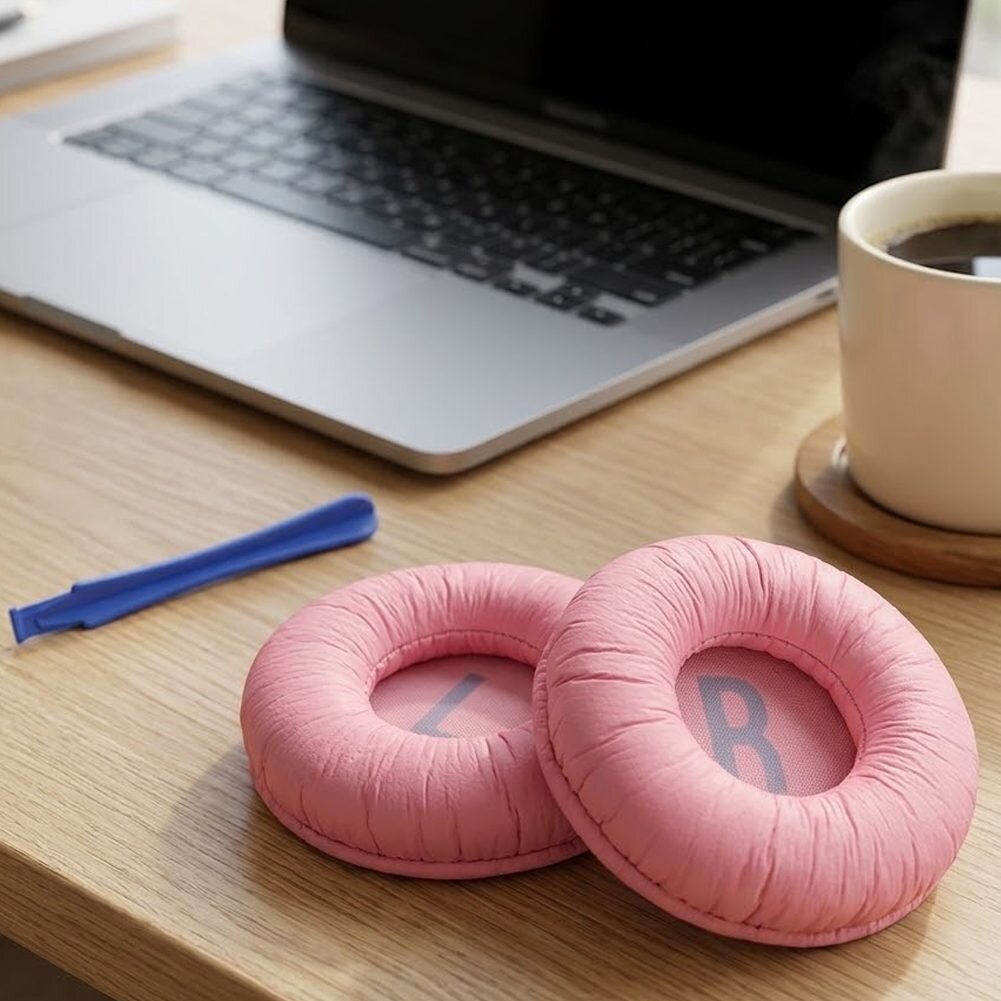 Na drewnianym biurku leżą różowe nauszniki z literami \'L\' i \'R\', obok niebieski plastikowy element, w tle otwarty laptop i filiżanka kawy.